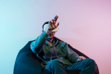 Pouf 'un üzerinde oturan ve görünmez nesnelere dokunan kapüşonlu genç bir erkek sanal dünyaya daldırılmış VR kulaklık kullanıyor.