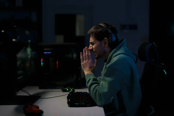 暗い部屋でコンピュータ上でビデオゲームをプレイしながら ヘッドセットの祈りで若いストレスの男性サイバースポーツゲーマーのサイドビュー — ストック写真
