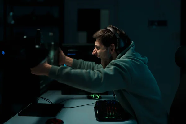 暗い部屋で成功したゲームを祝いながら ヘッドセットに触れるコンピュータモニターで男性のサイバースポーツゲーマーのサイドビュー — ストック写真