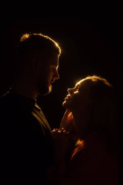 Genç sakallı adam ve sarışın kadının el ele tutuşup ışığa karşı karanlıkta nazikçe öpüşmesi.