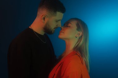 Mavi neon ışıkla karanlık odada dikilirken günlük kıyafetlerle birbirine dokunan genç adam ve kadının yan görüntüsü.