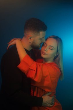 Mavi neon ışıkla karanlık odada dikilirken günlük kıyafetlerle birbirine dokunan genç adam ve kadının yan görüntüsü.