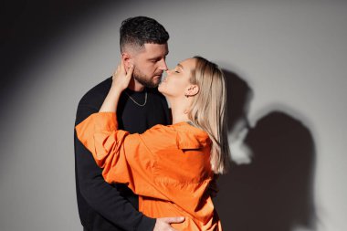 Side view of young man embracing and kissing girlfriend against wall with shadow from glowing light in dark studio clipart