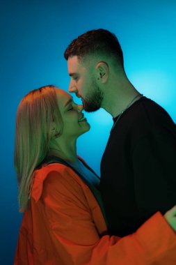 Mavi neon ışıkla karanlık odada dikilirken günlük kıyafetlerle birbirine dokunan genç adam ve kadının yan görüntüsü.