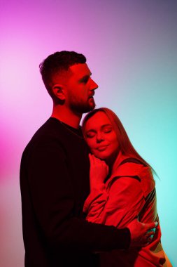 Genç sakallı adam renkli neon ışıkla stüdyoda dururken kız arkadaşına nazikçe sarılıyor.