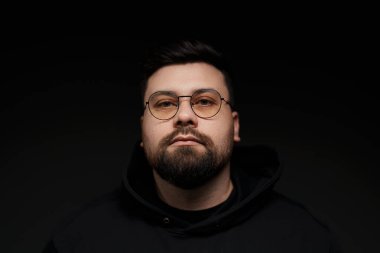 Gözlüklü, kapüşonlu, siyah arka planda kameraya bakan ciddi, sakallı, genç bir hippi.