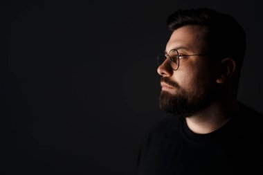 Moda güneş gözlüklü, duygusuz genç sakallı erkek model karanlıkta dikilirken düşünceli bakışlarını kaçırıyor.