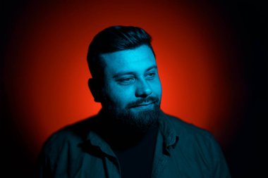 Tişörtlü, ciddi sakallı genç bir erkek model karanlıkta neon ışıkla dikilirken düşünceli bakışlarını kaçırıyor.