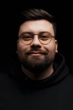 Gözlüklü, kapüşonlu, siyah arka planda kameraya bakan ciddi, sakallı, genç bir hippi.