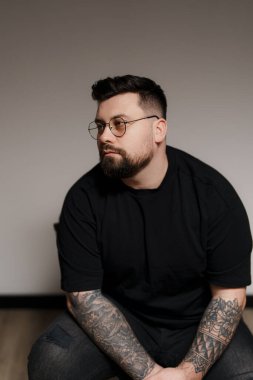Genç ve sakallı, siyah elbiseli, gözlüklü, kolları dövmeli, gri arka planda oturan genç bir erkek model.