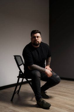 Genç ve sakallı, siyah elbiseli, gözlüklü, kolları dövmeli, gri arka planda oturan genç bir erkek model.