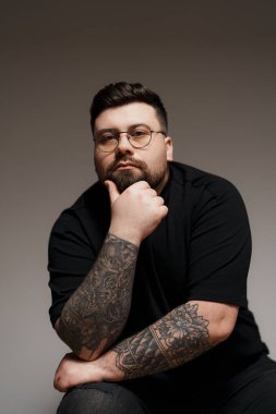 Genç ve sakallı, siyah elbiseli, gözlüklü, kolları dövmeli, gri arka planda oturan genç bir erkek model.