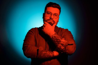 Tişört ve gözlük takmış, kırmızı ışıklı arka planda kameraya bakan, stüdyoda gölgesi olan duygusuz, sakalsız genç erkek modelin portresi.