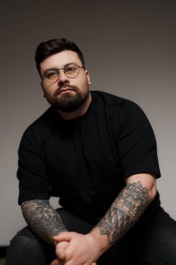 Genç ve sakallı, siyah elbiseli, gözlüklü, kolları dövmeli, gri arka planda oturan genç bir erkek model.