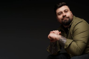 Yeşil tişörtlü, dövmeli, gözlüklü, gülümseyen ve stüdyoda sandalyede otururken kameraya bakan genç, pozitif sakallı bir erkek model.