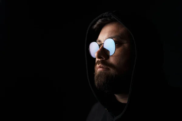 Unemotional young bearded male model in trendy sunglasses and black hood looking away thoughtfully while standing in darkness