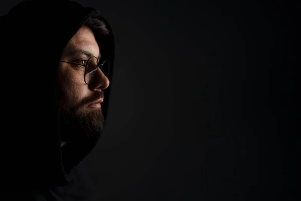 stock image Unemotional young bearded male model in trendy sunglasses and black hood looking away thoughtfully while standing in darkness