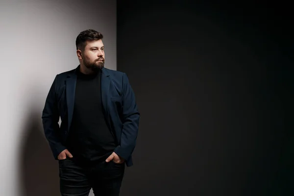 Pensive young bearded male model in elegant jacket and black pullover standing in studio with hands in pockets and looking away thoughtfully