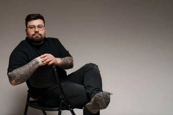 Content young plus size bearded male model in black outfit and eyeglasses with tattooed arms and crossed legs sitting on chair in studio