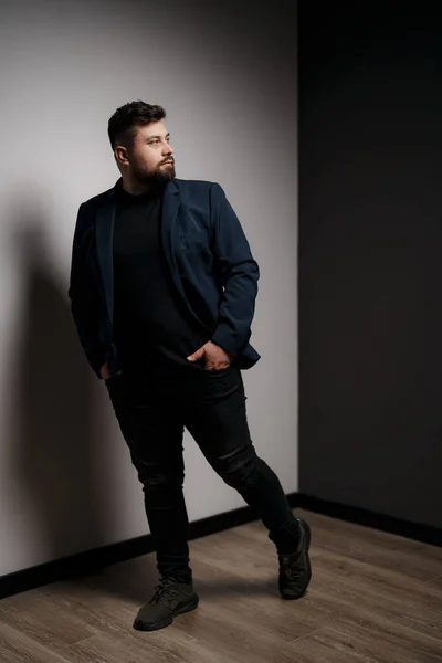 Pensive young bearded male model in elegant jacket and black pullover standing in studio with hands in pockets and looking away thoughtfully