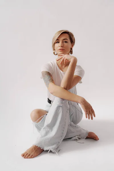 Full body of young barefoot female sitting on floor while relaxing against white background