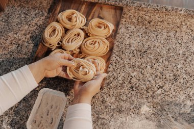 Yüksek açılı, isimsiz kadın aşçı. Mutfakta yemek hazırlarken, ahşap kesme tahtasına el yapımı rulo tagliatelle makarna koymuş.