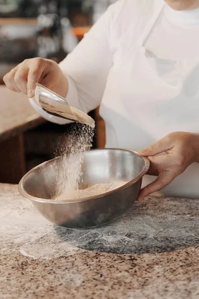 キッチンで生地を準備しながら 金属ボウルにスクープから小麦粉を注ぐ白いエプロンで匿名の女性料理をクロップ — ストック写真