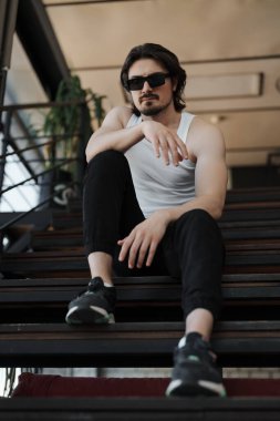 Düşük açılı, sakalsız, uzun siyah saçlı, sıradan elbiseli genç bir adam merdivenlerde oturmuş kameraya bakıyor.