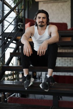 Düşük açılı, sakalsız, uzun siyah saçlı, sıradan elbiseli genç bir adam merdivenlerde oturmuş kameraya bakıyor.