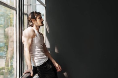 Beyaz kolsuz gömlekli, siyah pantolonlu, uzun saçlı kaslı bir adam panoramik pencerenin yanında duruyor ve başını çevirirken korkuluklara yaslanıyor.