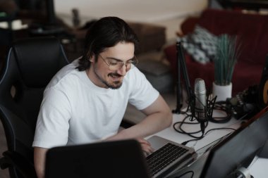 Gülümseyen sakallı, gözlüklü, beyaz tişörtlü bir müzisyen dizüstü bilgisayarla müzik bestelerken stüdyoda modern ekipmanlarla podcast yapıyor.