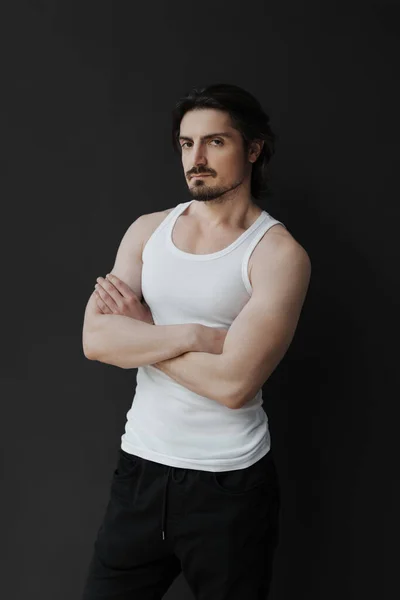 Self assured muscular bearded man with black hair in white sleeveless t shirt and pants standing on black background and looking at camera