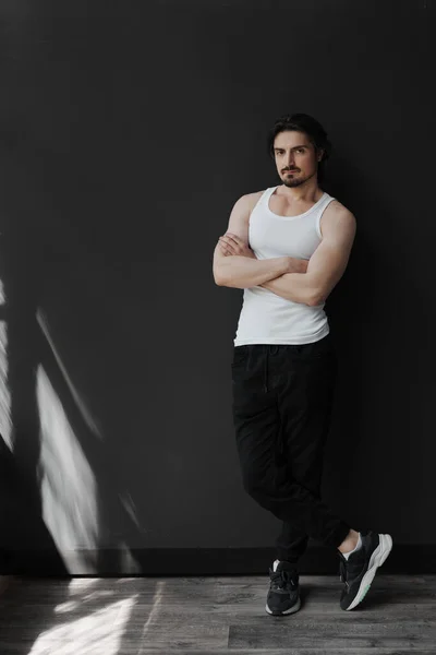 Self assured muscular bearded man with black hair in white sleeveless t shirt and pants standing on black background and looking at camera