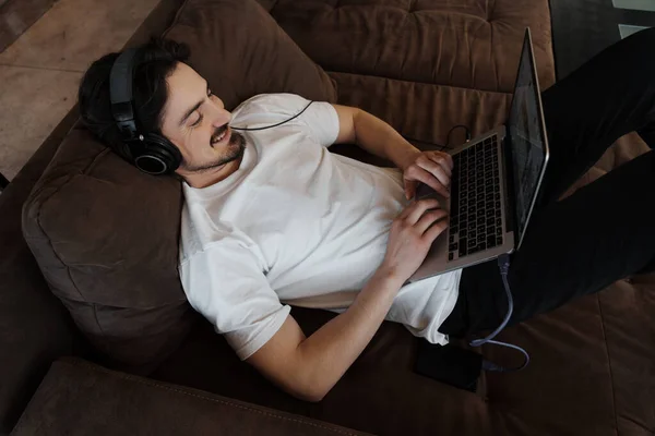 티셔츠와 헤드폰을 소파에 누워서 노트북을 음악을 작곡하는 남성의 위에서 — 스톡 사진