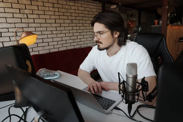 眼鏡と現代的な機器でスタジオでポッドキャストを記録しながら ラップトップ上で音楽を作曲白いTシャツで笑顔髭男性音楽家 — ストック写真