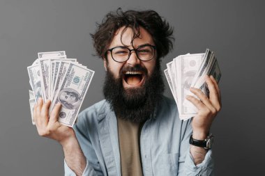 Yakışıklı sakallı adam elinde para tutuyor. Kot gömlekli memnun sakallı adam gri duvarda izole edilmiş bir tomar para gösteriyor..