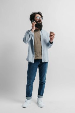 Yakışıklı bir hippinin uzun vücut portresi şık kıyafetler giymiş kahve içiyor ve beyaz arka planda akıllı telefondan konuşuyor.
