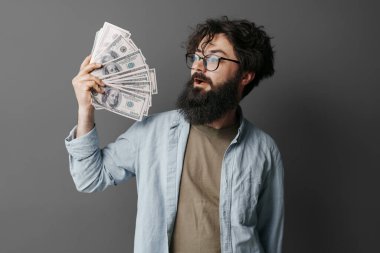 Yakışıklı sakallı adam elinde para tutuyor. Kot gömlekli memnun sakallı adam gri duvarda izole edilmiş bir tomar para gösteriyor..