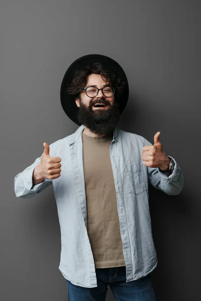 陽気な幸せな髭の男は眼鏡と黒の帽子を身に着けています 黒の背景に親指を表示 スタジオショット — ストック写真