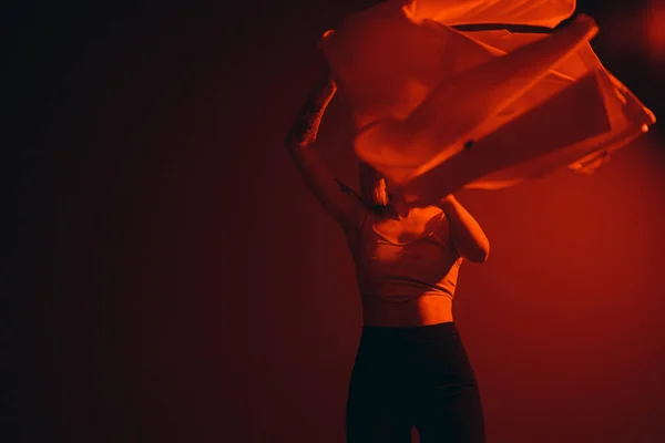 Lady in crop top raising trendy jacket against black backdrop in dim red light. Fashion concept