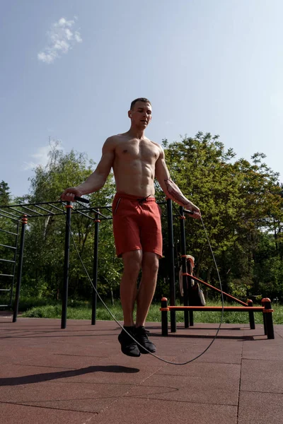 Full body of muscular shirtless male athlete working out on skipping rope during fitness training on sports ground during sunny summer day
