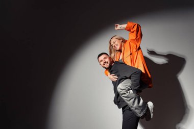 Siyah elbiseli neşeli bir adamın turuncu ceketli heyecanlı kız arkadaşını sırtında taşıması ve karanlık odada ışık altında elini kaldırması.