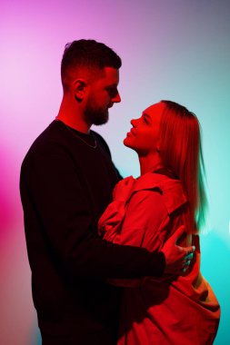 Stüdyoda renkli neon ışıklı arka planda dururken kız arkadaşına nazikçe sarılan genç sakallı adamın dikey fotoğrafı.