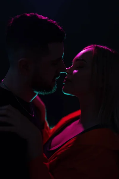 Side view of young couple kissing each other with closed eyes while standing in darkness with neon illumination