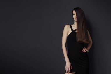 Siyah elbiseli, zayıf, genç bir kadın model ve uzun düz saçlı gri arka planda duruyor. Beline nazikçe dokunuyor. Uzun düz saçlı.