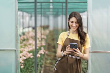 Smiling florist businesswoman using smartphone, wearing yellow t-shirt and brown apron holding phone in hands at the green house