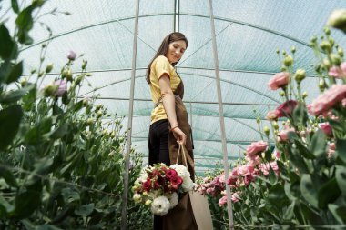 Woman florist walking among flowers in a green house carrying a basket with a fresh bouquet