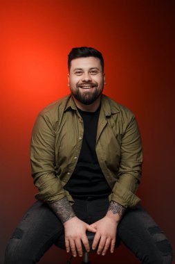 Pozitif, genç, sakallı, gündelik elbiseli erkek model. Parlak kırmızı arka planda dişlek bir gülümsemeyle sandalyede oturuyor.