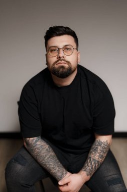 Siyah tişörtlü ve yırtık kotlu bir adam, taburede oturuyor. İki kolunda da dövmeleri var. Arka plan düz beyaz bir duvar.