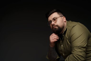 Gözlüklü, yeşil gömlek giyen düşünceli sakallı bir adamın portresi. O, çenesini elinin üzerinde dinlendiriyor ve kameradan uzağa bakıyor. Arka plan koyu gri.